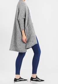 Harmaa ylisuuri neuletakki, joka on valmistettu pehmeästä neuloksesta ja jossa on pitkät hihat, etutaskut sekä avoin etuosa. Se on yhdistetty sinisiin farkkuihin ja mustiin lenkkareihin.