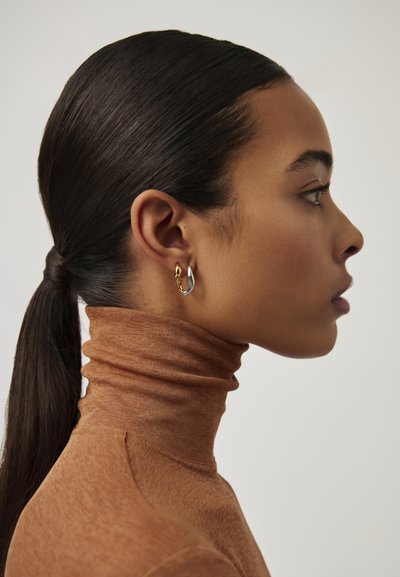 Χρυσά και ασημένια σκουλαρίκια κρίκοι σε μια μοντέλο, που φοράει μια καφετί ζιβάγκο με υφή. Τα μαλλιά είναι στιλιζαρισμένα σε μια κομψή αλογοουρά.