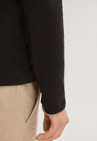 Schwarzes Langarmhemd mit glatter Textur, abgerundetem Saum und tailliertem Bündchen, kombiniert mit hellbeigen Hosen.