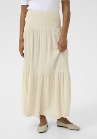Light beige maxi skirt with a smocked waistband and tiered design, made from textured fabric. Paired with beige flat shoes.