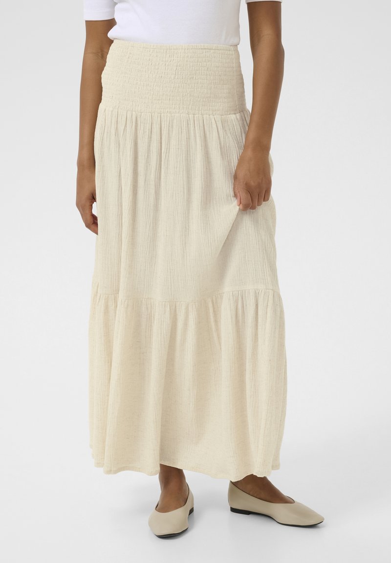 Light beige maxi skirt with a smocked waistband and tiered design, made from textured fabric. Paired with beige flat shoes.