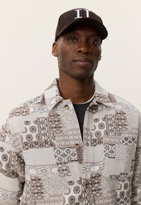 Πουκάμισο με κουμπιά στο εμπρόσθιο μέρος σε ανοιχτό μπεζ χρώμα με περίτεχνα μοτίβα από καφέ και μαύρα πατς, με διακοσμημένο γιακά τύπου spread και δύο κουμπιά. Καπέλο καφέ χρώματος πάνω από το πουκάμισο.