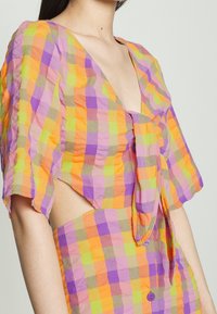 Blouse à carreaux colorée en violet, orange et vert. Manches courtes et larges, décolleté en V profond, avec un détail noué à l'avant. Tissu léger et texturé.