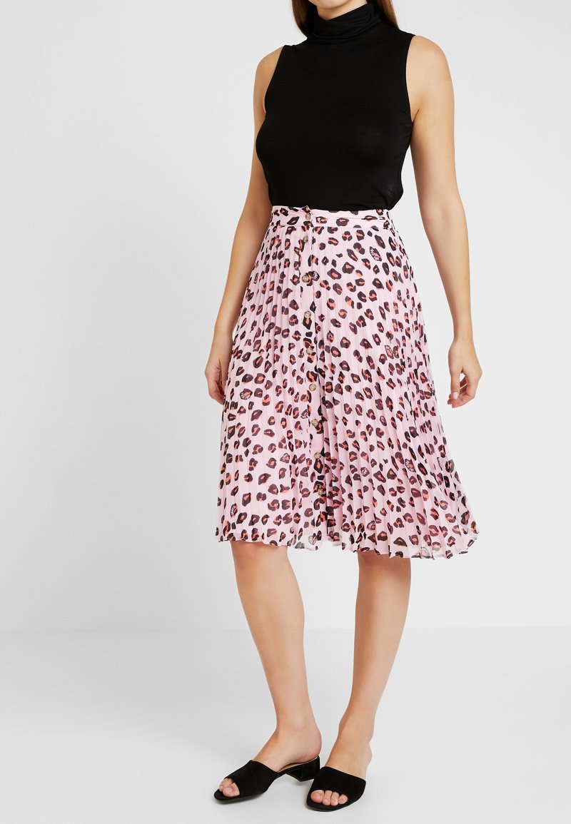 Femme portant un haut noir sans manches à col roulé, une jupe midi plissée rose à imprimé léopard avec une patte de boutonnage sur le devant, assortie à des talons ouverts noirs.