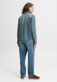 Giacca in denim blu chiaro e jeans morbidi, con yoke posteriore, polsini con bottoni e leggere sfumature, abbinati a scarpe scure.