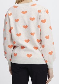 Pull beige clair avec motifs de cœurs orange, col rond et poignets côtelés, manches ballon et finition en maille texturée.