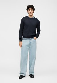 Jeune homme debout, une main dans la poche, portant un pull noir, un jean bleu clair à jambes larges et des chaussures noires sur un fond gris uni.