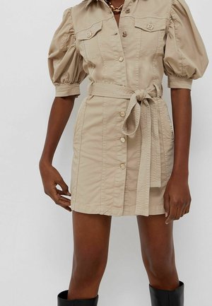 Robe beige boutonnée avec manches bouffantes et ceinture nouée à la taille, portée avec un collier chaîne doré et des bottes noires montantes.
