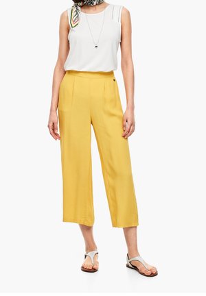 Donna che indossa pantaloni gialli a gamba larga e corti, top bianco senza maniche, sandali argento, collana e sciarpa a righe al collo su sfondo bianco.