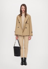 Manteau trench beige à double boutonnage, pull en maille, pantalon beige ajusté, sac seau noir avec des accents dorés et bottines noires à talons pointus.