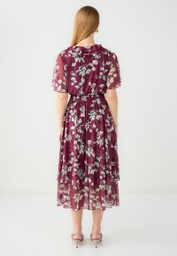 Mauve bloemdress met korte mouwen, elastische taille, gelaagde rok en doorzichtige stof met een gestructureerde afwerking. Zilveren hakken zijn zichtbaar.