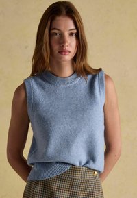 Noor naine, kellel on heledat pruunid juuksed, kannab ilma varrukateta helesinist kudumit ja kõrge vöökohtadega houndstooth seelikut, millel on kuldne nupp.