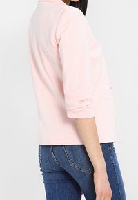 Světle růžový blazer s klopovým límcem, tříčtvrtečními rukávy a elegantním designem. Nosit s modrými džíny, obsahuje minimální švy.