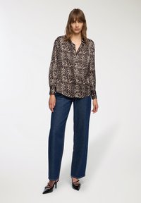 Kobieta w bluzie z długim rękawem w panterkę, ciemnoniebieskich dżinsach o szerokich nogawkach i czarnych szpilkach na tle jednolitego tła.