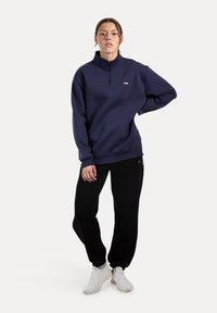 Mujer de pie con una mano en la cadera, vistiendo un pulóver azul marino de media cremallera, pantalones jogger negros y zapatillas blancas, sobre un fondo blanco.