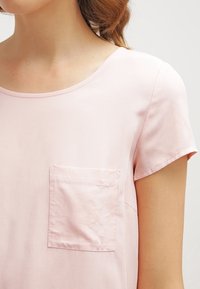 Světle růžový top s krátkým rukávem vyrobený z hladké látky, s kulatým výstřihem a obdélníkovou kapsou na levé straně.