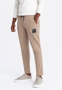 Beige joggerbyxor i mjukt material, slim fit, med en svart logotyp på låret, och matchade med ljusgrå sneakers.