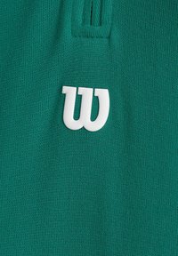 Tessuto verde con una superficie texturizzata, con un prominente logo bianco a forma di "W" come dettaglio distintivo. Il materiale appare morbido e lavorato a maglia.