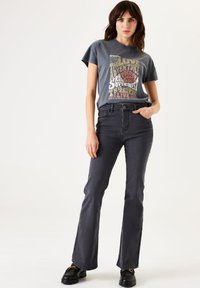 Camiseta gráfica gris con texto colorido, combinada con pantalones de mezclilla acampanados gris oscuro. El modelo lleva zapatos negros gruesos. Fondo simple.