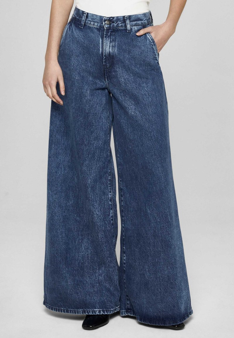 Mėlynos plačios džinsinės kelnės su išblukusiu atspalviu, aukštu liemeniu, priekinėmis kišenėmis ir klasikiniu užsegimu su saga. Kelnės apačia baigiama šiek tiek trupančiu apvadu.