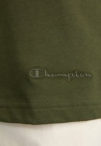 Olive green cotton t-shirt with ribbed texture, featuring a stitched "Champion" logo in tonal thread near the hem. Simple, crew neck design.