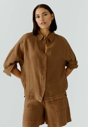 Frau mit dunklem Haar, die ein lockeres braunes Leinenhemd mit Knöpfen und passende Shorts trägt, steht mit den Händen hinter dem Rücken vor einfarbigem Hintergrund.