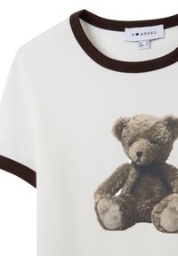 Camiseta de algodón blanca que presenta un oso de peluche marrón impreso con una cinta negra. La camiseta tiene un cuello redondo de canalé marrón.