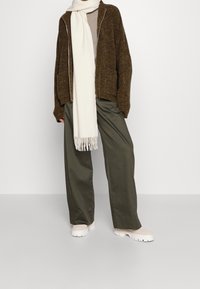 Brauner gestrickter Cardigan mit einem Reißverschluss, beiger Schal mit Fransen, beige Schuhe und olivgrüne weit geschnittene Hosen. Minimalistisches Design und sanfte Texturen.