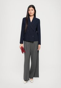 Blazer blu navy doppiopetto con una vestibilità su misura, abbinato a pantaloni grigi a gamba larga. La modella tiene una pochette rossa e indossa scarpe color crema.