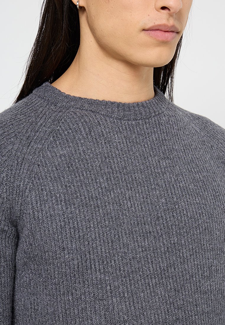 Primo piano di una persona che indossa un maglione grigio lavorato a maglia, che mostra il collo, la spalla e parte del viso con capelli lunghi e scuri e un piccolo orecchino a cerchio.