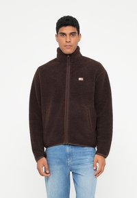 Jachetă din fleece maro, cu guler înalt, fermoar frontal și buzunare laterale. Prezintă un material texturat și un mic logo de marcă pe piept.