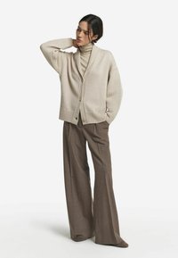 Cardigan en tricot beige avec décolleté en V, coupe ample et deux poches avant, associé à un pantalon large marron. Texture douce, détails minimalistes.