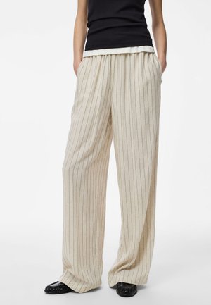 Personne portant un pantalon beige à fines rayures larges, un haut noir et des chaussures noires faciles à enfiler, debout les mains dans les poches sur un fond blanc.