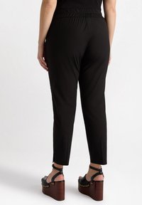 Zwarte elastische broek met een taps toelopende fit en zijsplitsen, gecombineerd met zwarte wedge sandalen met een houten plateau en gouden studs.