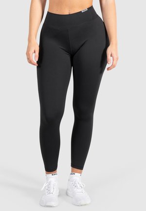 Leggings noirs avec une taille haute, texture lisse et design moulant, associés à des chaussures de sport blanches. Pas de motifs distincts.