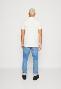 Camiseta blanca de manga corta con un corte recto, combinada con unos jeans azul claro y zapatos negros. La vista desde atrás muestra el diseño limpio de la camiseta.