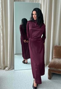 Donna in un lungo abito bordeaux e tacchi neri sta in piedi con le mani incrociate dietro, riflessa in uno specchio a figura intera in una stanza con tende beige.