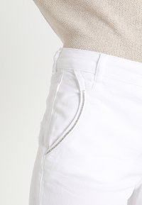 Pantalones de algodón blanco con mezcla, que presentan un corte slim, costuras decorativas plateadas a lo largo de los bolsillos y una cintura de altura media. Parte superior de punto texturizado visible.