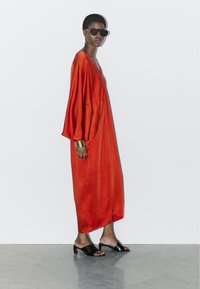 Lange, vloeiende rode jurk met brede mouwen, gemaakt van gladde stof. Gecombineerd met zwarte mules en donkere zonnebril, met een metalen armband.