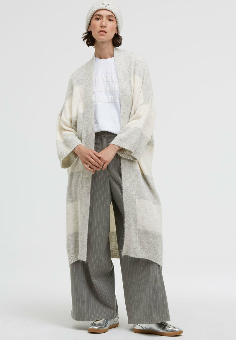 Light gray and cream patterned oversized cardigan with wide sleeves, layered over a white shirt and gray striped trousers, paired with silver shoes.