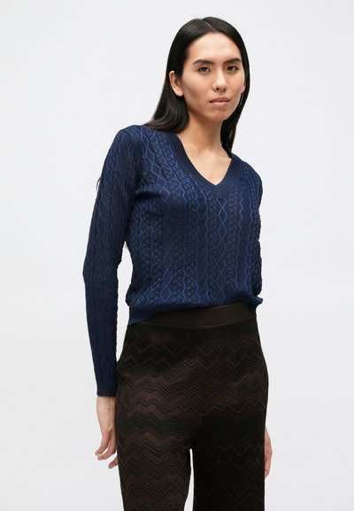 Tamsiai mėlynas V formos kaklo megztinis su tekstūruotu geometriniu raštu; lengva mezgimo medžiaga. Derinamas su tamsiai rudos spalvos kelnėmis su chevrono raštu.