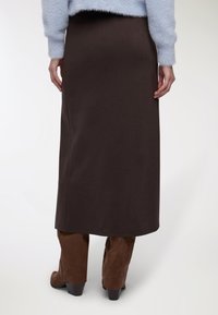 Persona che indossa una gonna midi marrone scuro, un maglione fuzzy azzurro chiaro e stivali con tacco in suede marrone, posando su uno sfondo neutro.