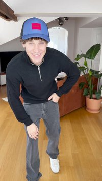 Black knitted sweater with a zip collar, grey jeans, and white sneakers. A blue cap with a red "HUGO" logo completes the outfit. Indoor setting.