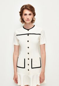 Cremefarbenes Kleid mit kurzen Ärmeln, runder Halslinie, schwarzen Akzenten, vorderem Knopfverschluss und zwei aufgesetzten Taschen. Ausgestellter Saum.