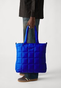 Sac fourre-tout bleu rembourré avec une texture matelassée, doté de deux poignées solides et d'un logo subtil à l'avant. Personne tenant le sac.