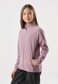 Jauna moteris dėvi šviesiai violetinį švarkelį su užtrauktuku, rankomis kišenėse, stovinti priešais baltą foną.