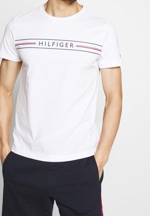 Mand iført en hvid T-shirt med korte ærmer med teksten "HILFIGER" og stribet design, kombineret med mørke bukser, hænderne i lommerne.