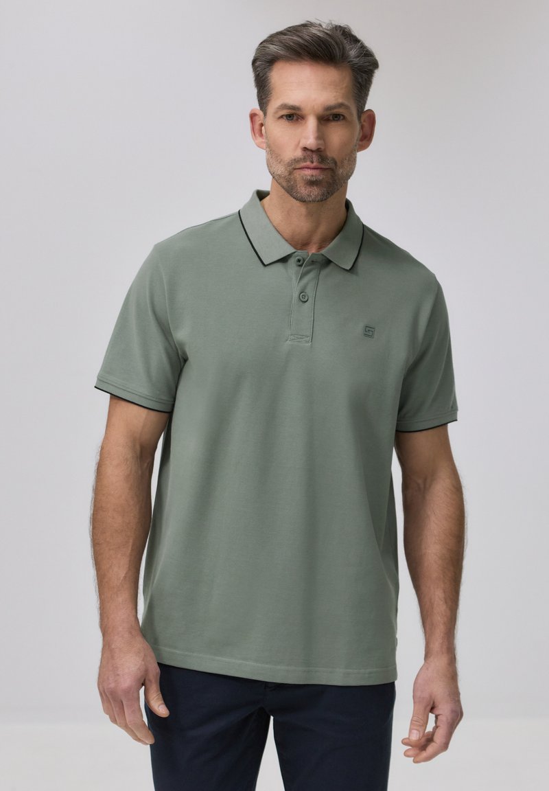 Homme portant un polo à manches courtes vert sauge avec des bordures noires sur le col et les manches, associé à un pantalon foncé, debout devant un fond uni.