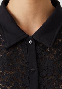 Svart spetstopp med klassisk krage, texturerat blommönster och knäppning vid nacken med svarta knappar.