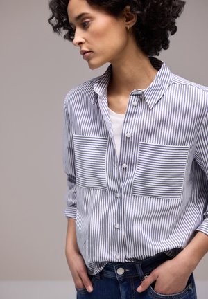 Kvinde med kort krøllet hår iført en stribet skjorte over en hvid top, poserende med hænderne i lommerne på sine cowboybukser.